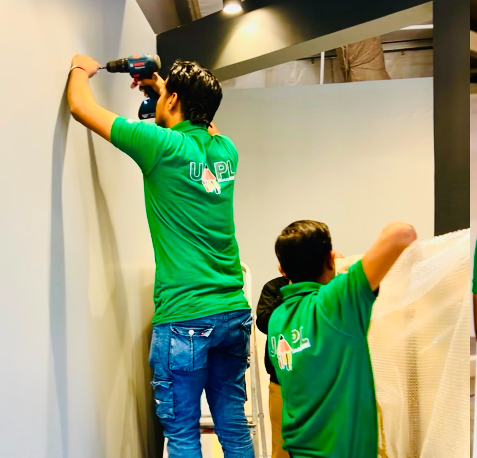 A UAPL team member on a ladder drilling into a gallery wall while a colleague holds bubble wrap